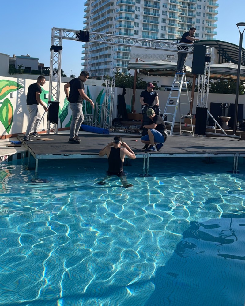 Concert Stage Production Setup Music To Go crew assembling a professional concert stage and lighting truss system over a swimming pool for an outdoor corporate event.
