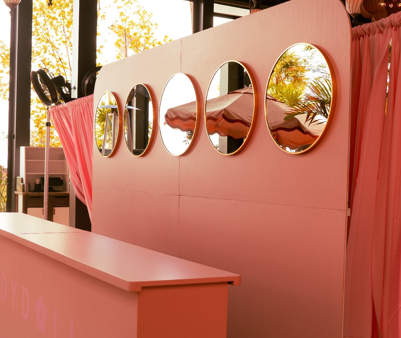 Custom Event Production Vanity Wall Mirror Rental Premium outdoor event brand activation setup featuring a custom-built pink partitions wall with integrated gold-trimmed oval vanity mirrors, a sleek matching console bar structure, and elegant drapery for luxury experiential marketing productions.