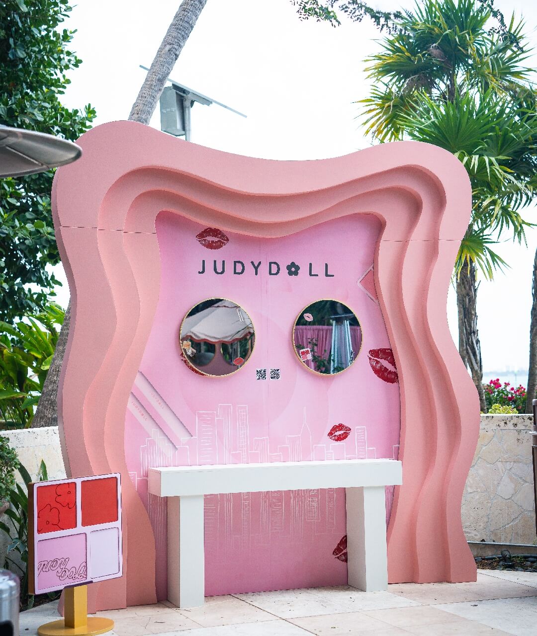 Custom Fabricated Brand Activation Vanity Backdrop Custom-designed outdoor brand activation booth featuring a layered wavy pink structure, integrated round vanity mirrors, a sleek console table, and custom cosmetics props, highlighting premium experiential marketing production and custom scenic fabrication rentals.